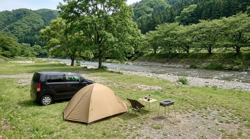 芝生と木陰のある河原の、川から離れた場所に車を横づけして張ったテントとバーベキューの道具(イメージ)